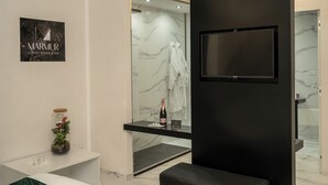 Um banheiro moderno com parede de mármore, televisão montada, box de vidro e uma garrafa de champanhe em uma toucador.