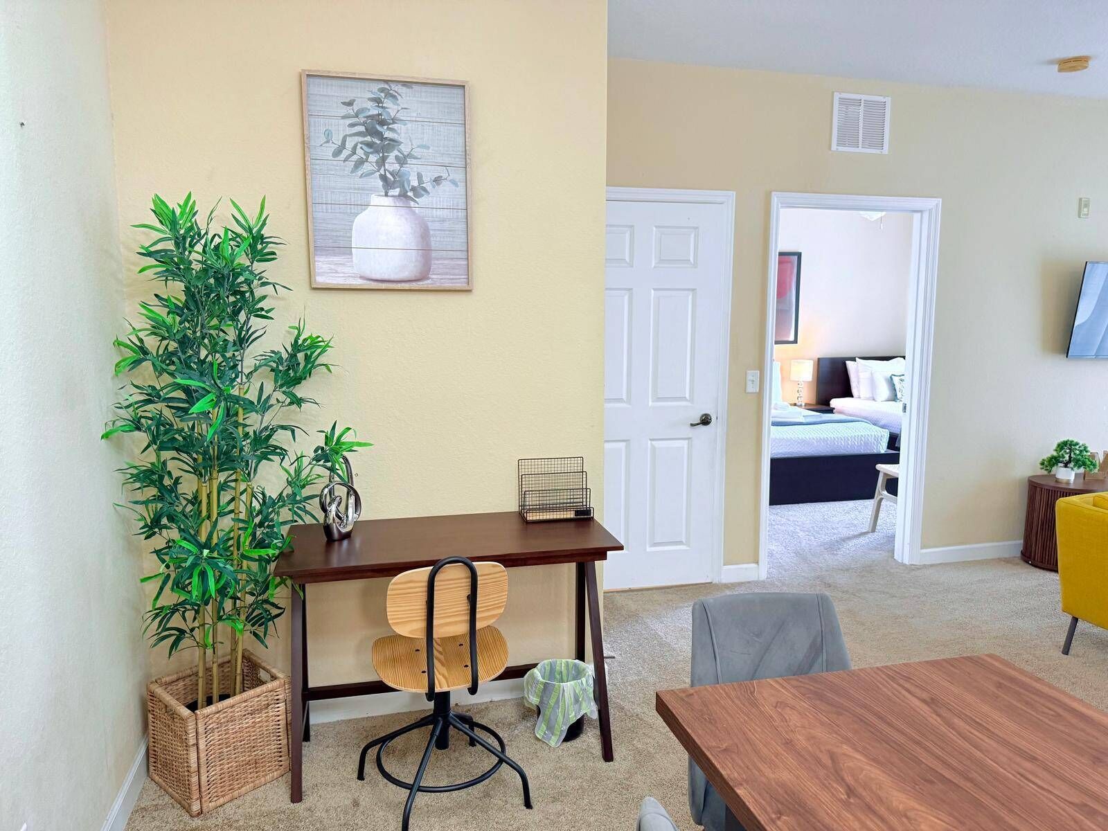 Una habitación con un escritorio de madera, una silla, una planta en maceta, un cuadro en la pared y una cama al fondo.