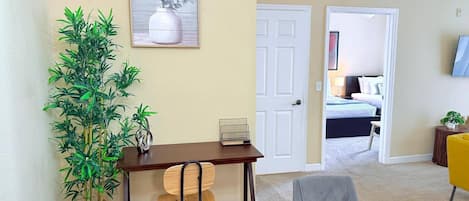 Una habitación con un escritorio de madera, una silla, una planta en maceta, un cuadro en la pared y una cama al fondo.