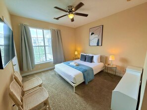 A hotel room with a bed, two bedside tables, a ceiling fan, a window with curtains, a chair, and a wall-mounted TV.