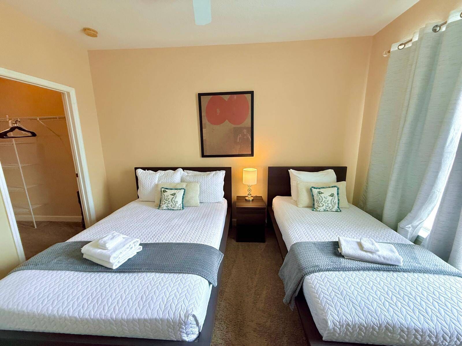 A hotel room with two beds, a closet, a painting on the wall, and a nightstand with a lamp.