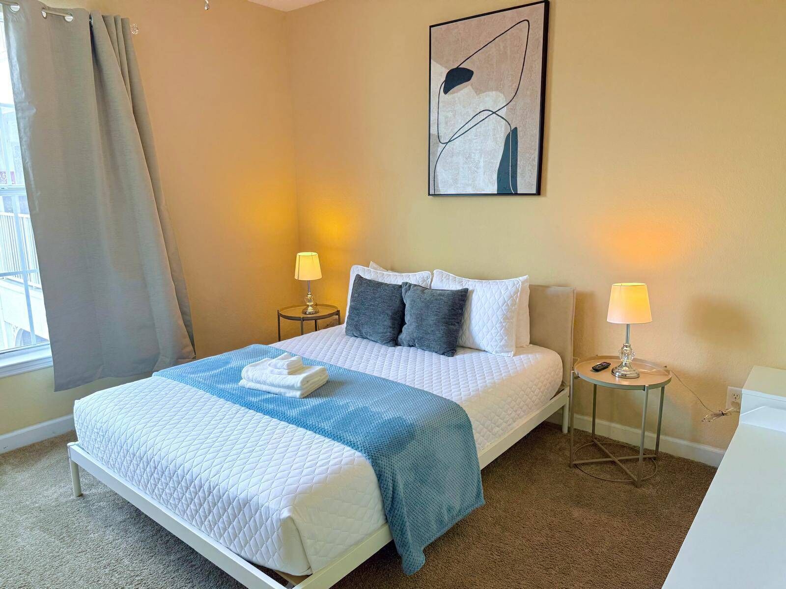 A hotel room with a bed, two bedside tables, a lamp, a painting, and a window with curtains.