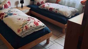 A room with two single beds, a nightstand with a lamp, a framed certificate on the wall, and a window with curtains. - Vacation apartment - Klenk - 1 (Gruibingen)