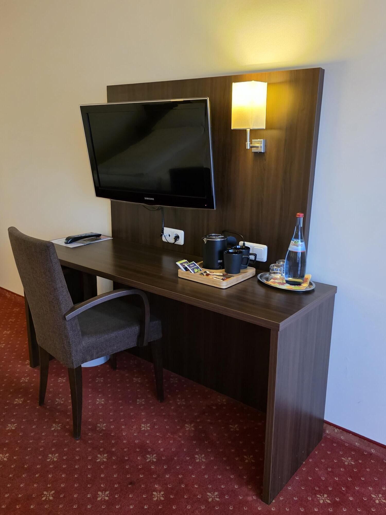 A hotel room with a wooden desk, a television, a chair, a remote control, a tray with a coffee pot, cups, and a bottle.