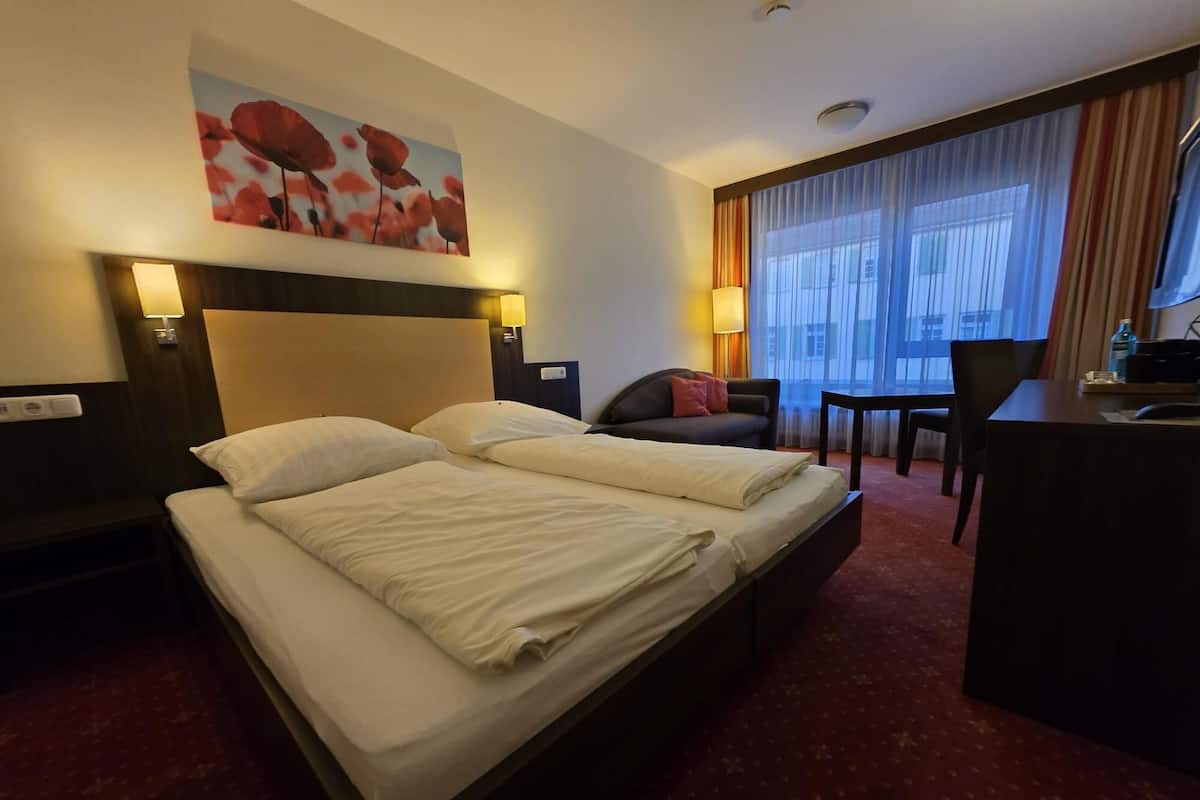 A hotel room with a large bed, a desk, a chair, and a large window with curtains.