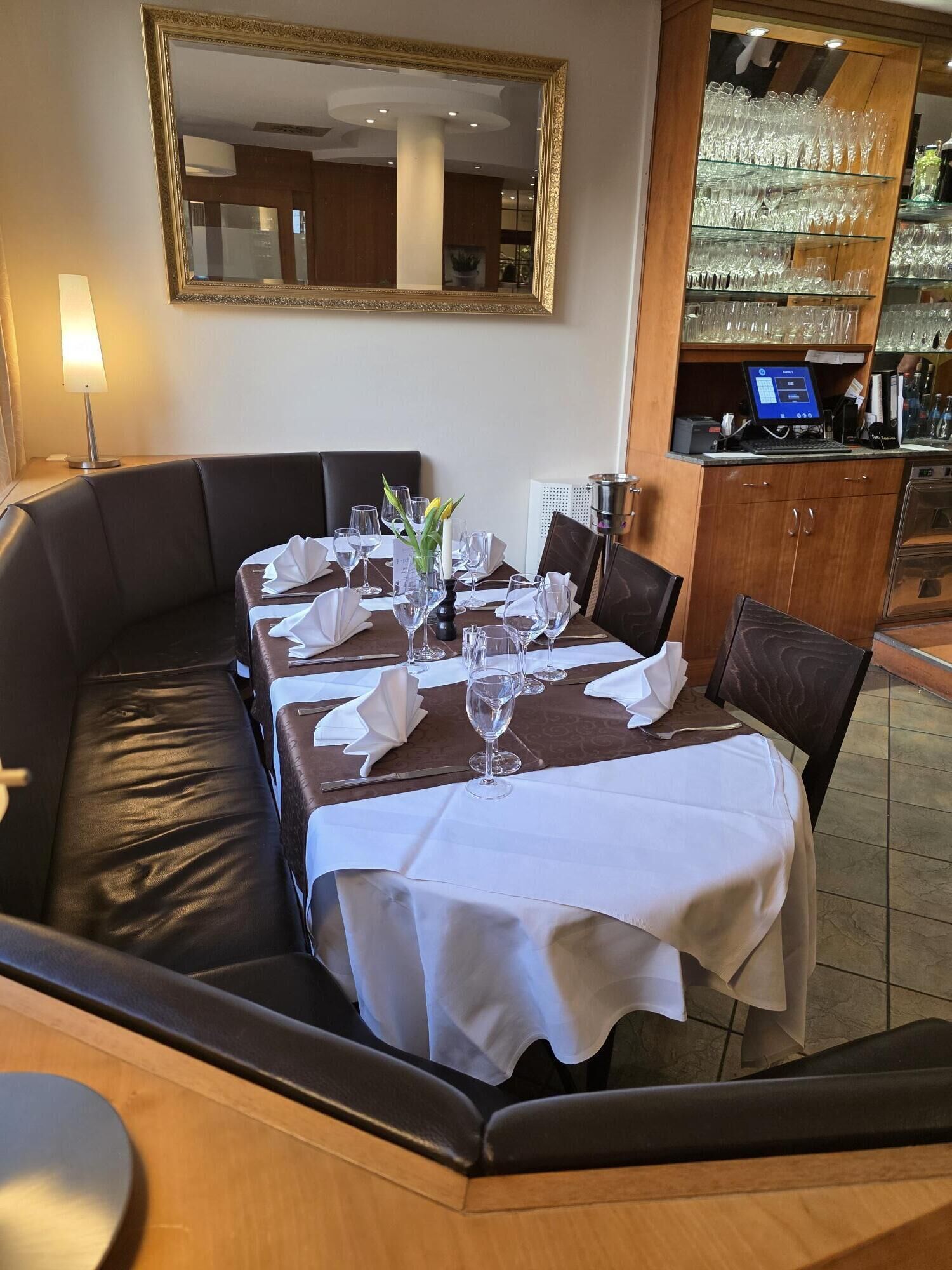 Ein Restaurant mit einem langen Esstisch, gedeckten Plätzen, einer Sofaecke und einer Bar mit einem Computer.
