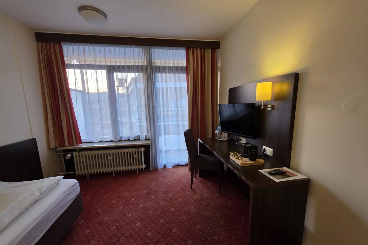 A hotel room with a bed, a desk with a computer, a TV, and a window with curtains.