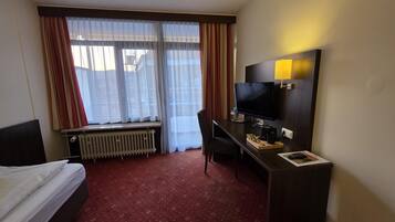 A hotel room with a bed, a desk with a computer, a TV, and a window with curtains.