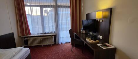 Une chambre d’hôtel avec un lit, un bureau avec un ordinateur, une télévision et une fenêtre avec des rideaux.