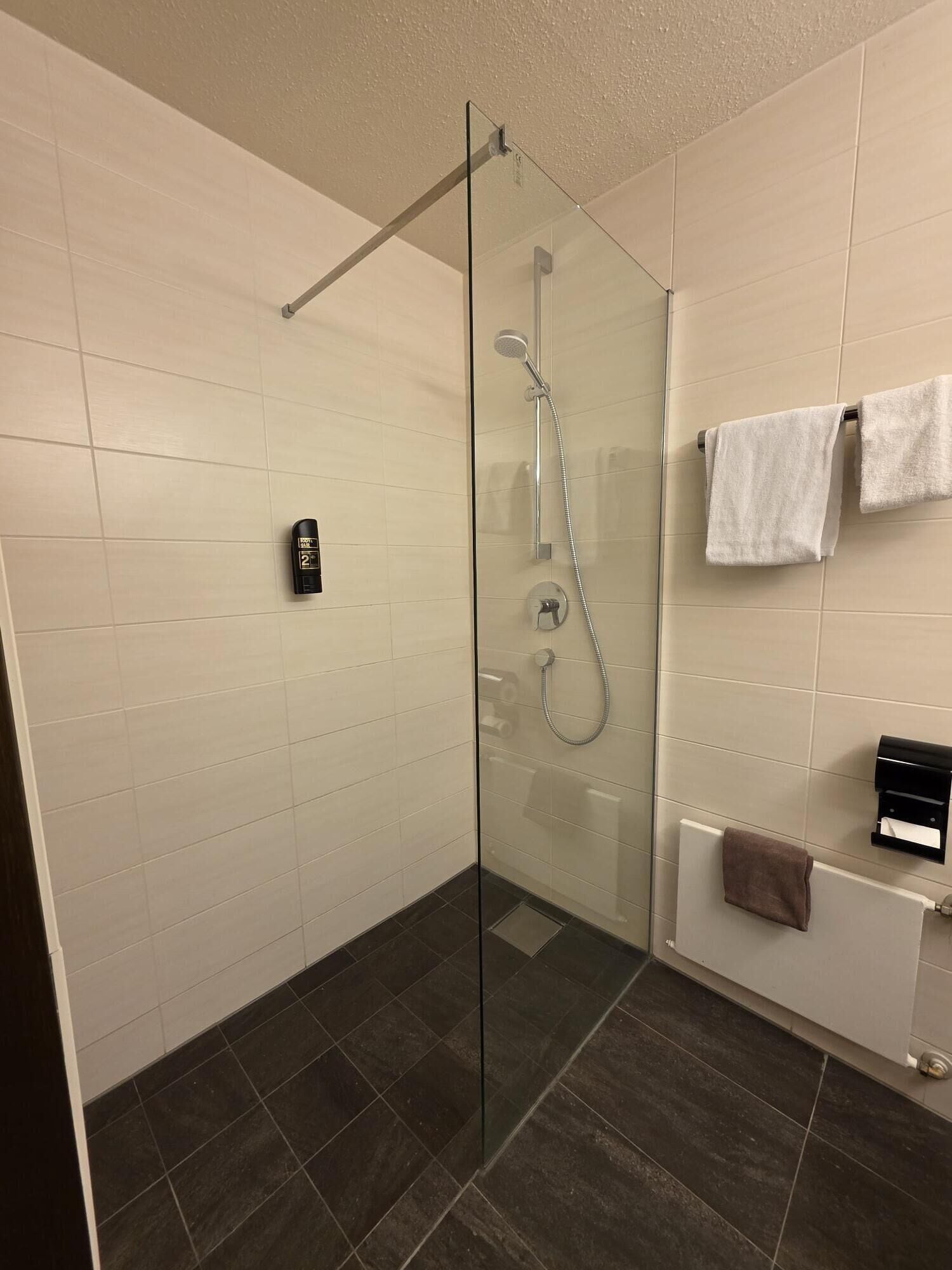 A modern bathroom with a glass-enclosed shower, white tiled walls, a dark floor, and a towel rack with two towels.