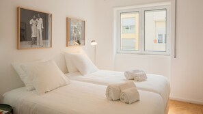 Habitación de hotel con dos camas, ropa de cama blanca, una lámpara de pie y dos fotografías enmarcadas en la pared.