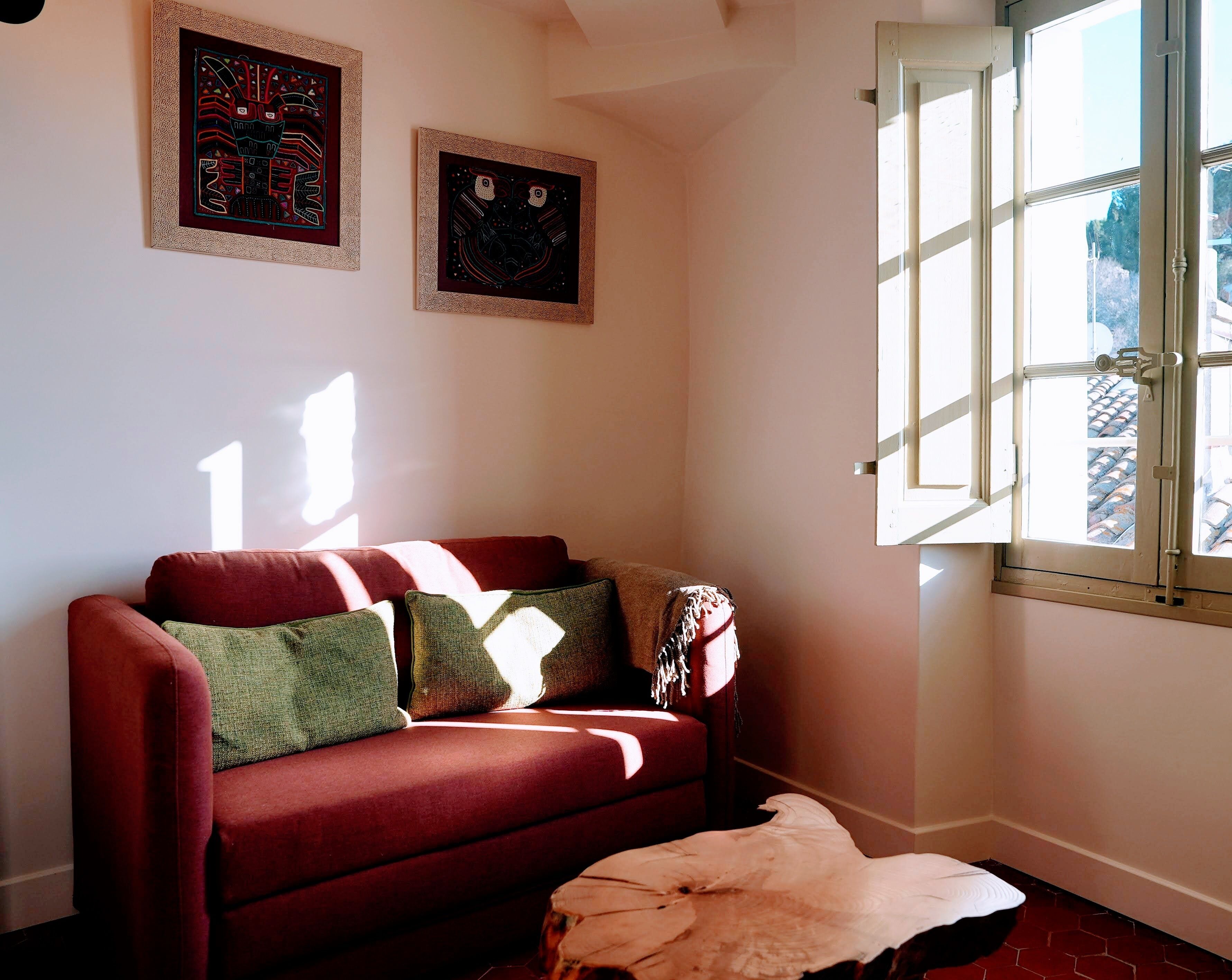 A cozy room with a red sofa, two framed artworks on the wall, and a large window with a view of the outside.