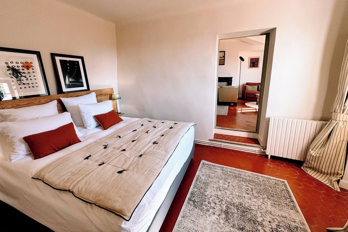 Una habitación de hotel con una cama grande, una mesita de noche con lámpara, una alfombra estampada en rojo y blanco, y vistas al pasillo y otra habitación.