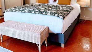 Un dormitorio con una cama, dos almohadas, una mesita de noche, una lámpara, un banco y dos cuadros enmarcados en la pared.