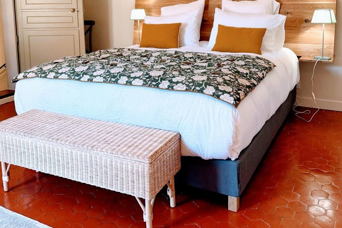 Un dormitorio con una cama, dos almohadas, una mesita de noche, una lámpara, un banco y dos cuadros enmarcados en la pared.