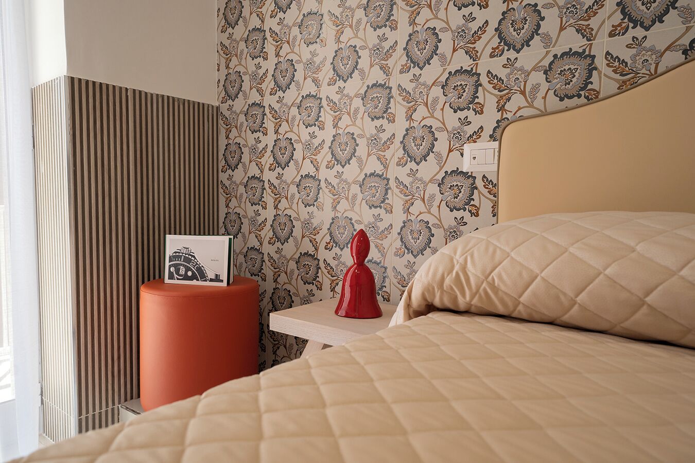 A hotel room with a bed, a nightstand with a red decorative item, and a striped wall.