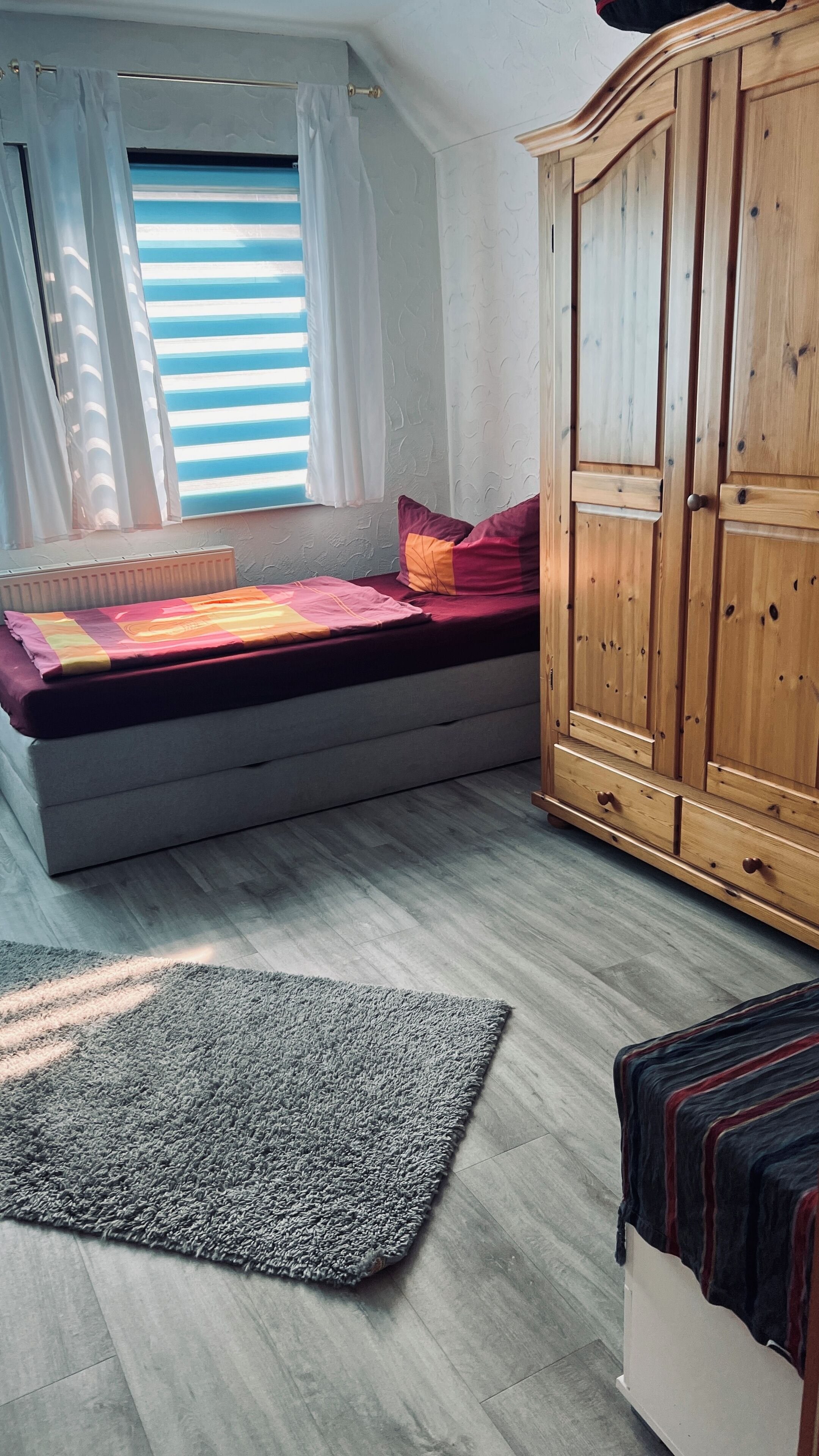 Een slaapkamer met een bed, een kledingkast, een raam met gordijnen en een grijs vloerkleed.