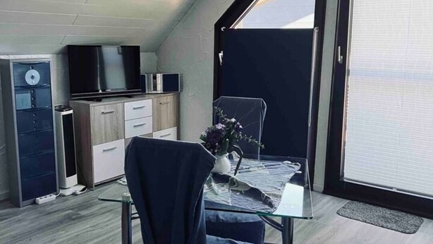 Une pièce de taille modeste, comprenant une table en verre, une chaise, un petit meuble, un téléviseur et une fenêtre avec des stores.