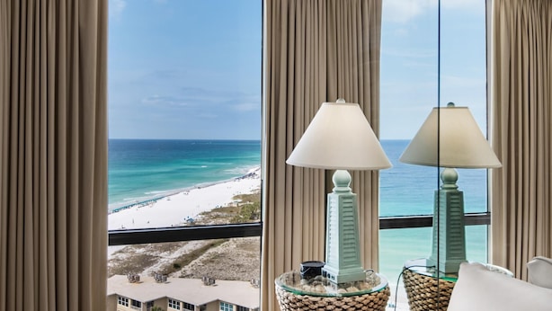 A room with a view of a beach and ocean. There are two lamps on a table.