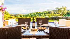 Une table à manger dressée pour quatre personnes, avec une bouteille de vin, des verres et des assiettes remplies de nourriture, donnant sur un paysage luxuriant.