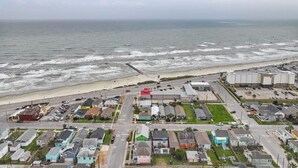 An aerial view of a coastal residential area with houses, a beach, and a road leading to a large industrial building. - High Tide of Your Life | Near Beach | Attractions (Galveston)