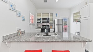 A modern kitchen with white cabinetry, a stainless steel refrigerator, granite countertops, and red bar stools. - High Tide of Your Life | Near Beach | Attractions (Galveston)