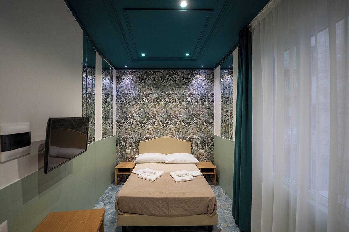 A narrow hotel room with a bed, a small table, and a mirror.