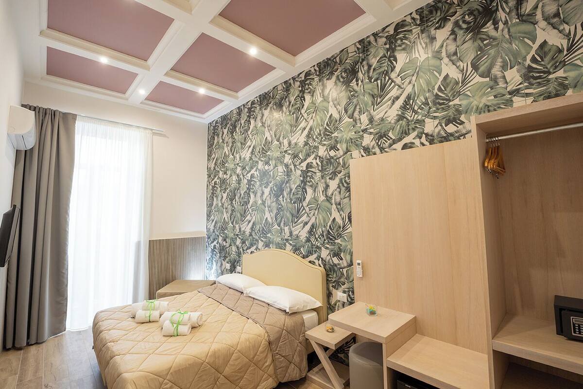 A hotel room with a bed, a bedside table, a wooden wardrobe, and a tropical leaf-patterned wall.