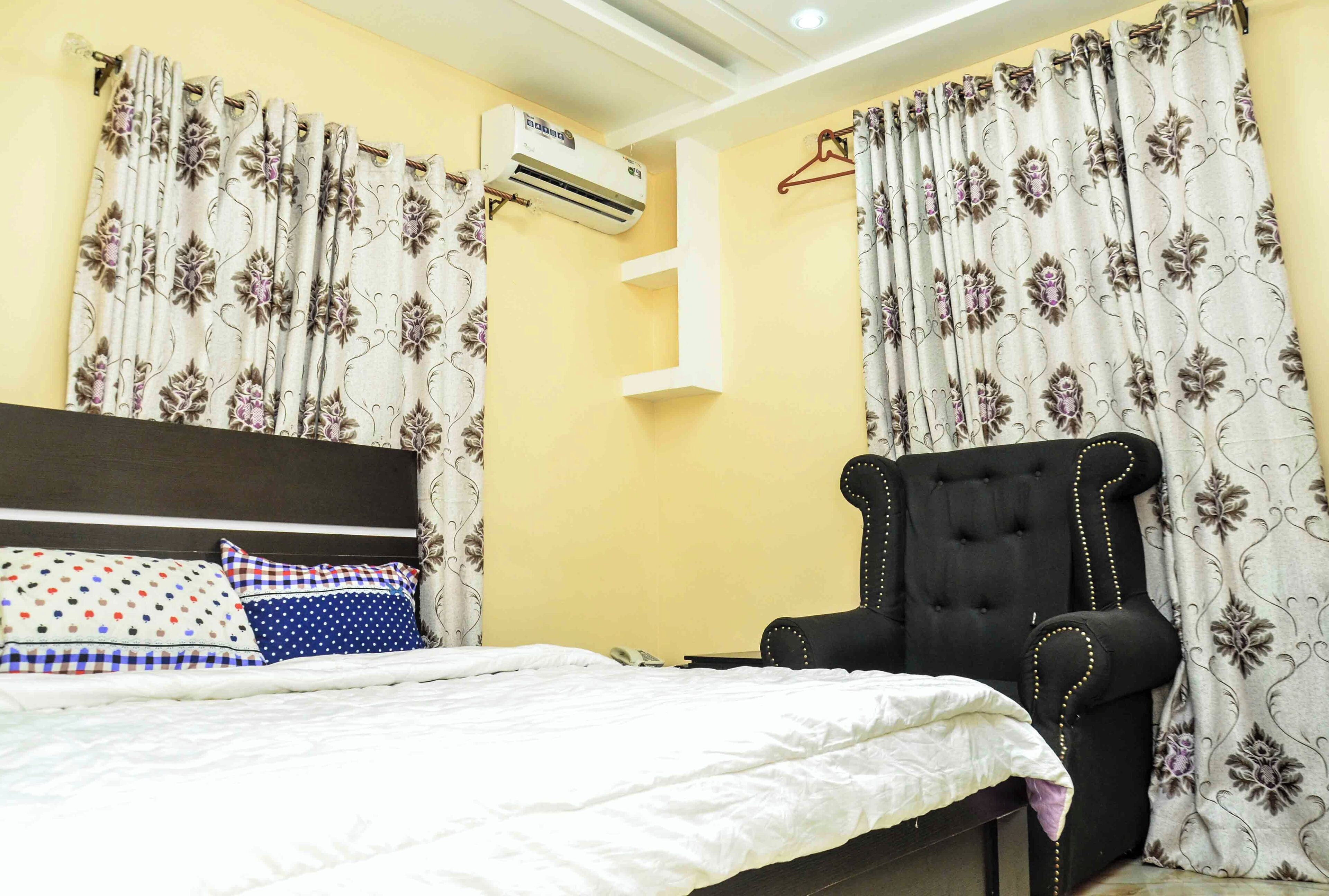 Een slaapkamer met een bed, een zwarte beklede fauteuil, bloemen gordijnen en een airconditioning.