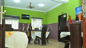 Un coin repas avec des tables disposées pour accueillir quatre personnes, un ventilateur de plafond, un téléviseur fixé au mur et une porte portant l’inscription « CUISINE ».