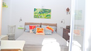 A bedroom with a bed, pillows, a chair, a bedside table, and a wall-mounted painting. - Double Room. The Garden. Rooms & Breakfast. (La Maddalena)