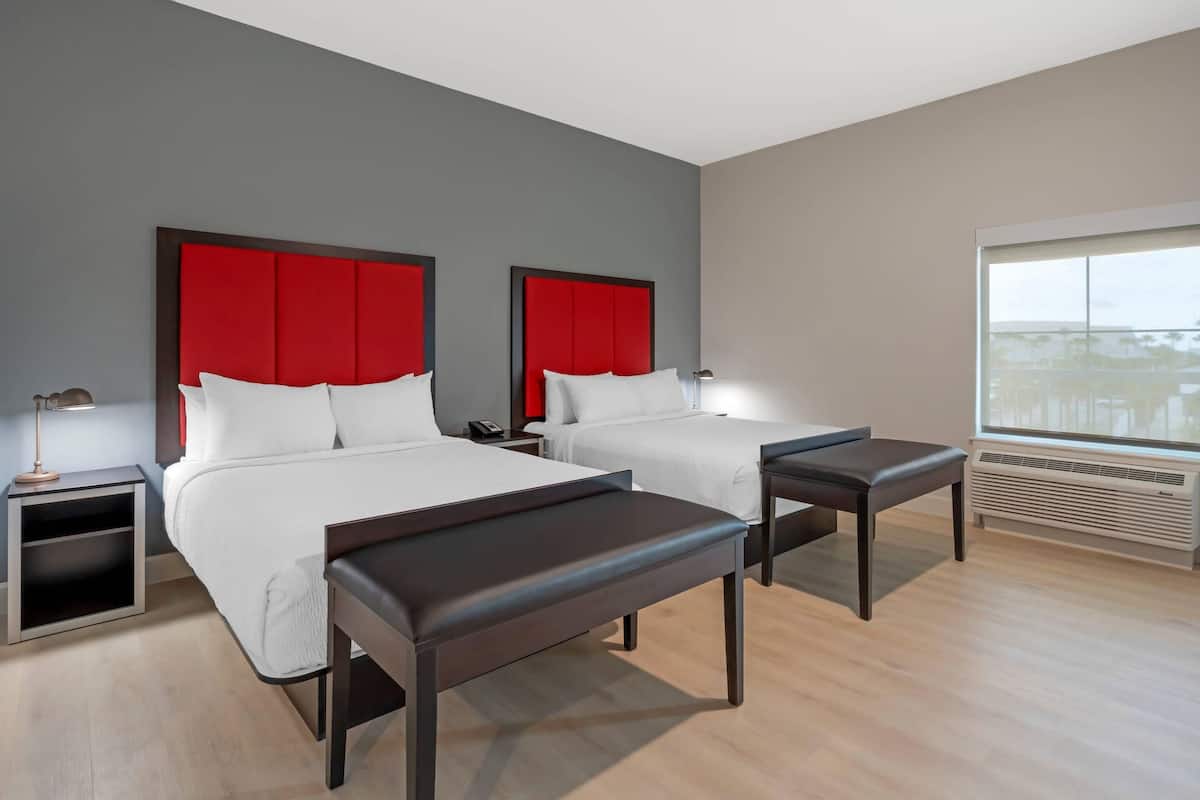Hotelkamer met twee bedden, een nachtkastje met lamp, een bankje, een raam met uitzicht en een airconditioning.