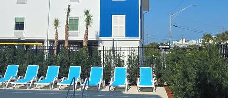 A modern hotel with a pool area, featuring blue and white exterior walls, a clear sky, and a row of blue lounge chairs.