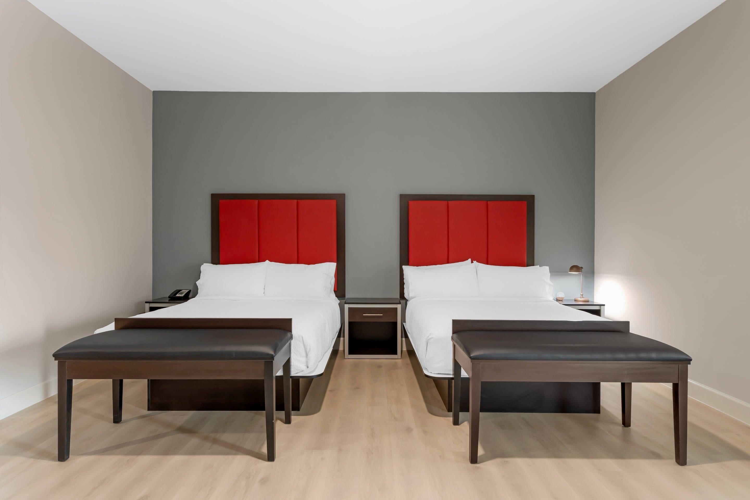 Quarto de hotel com duas camas, um banco, uma mesa de cabeceira e uma mesinha de apoio.