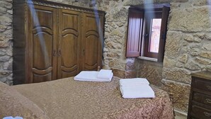 A room with a bed, two towels, a wooden wardrobe, a stone wall, and a window. - Village of Moura Morta (Castro Daire)