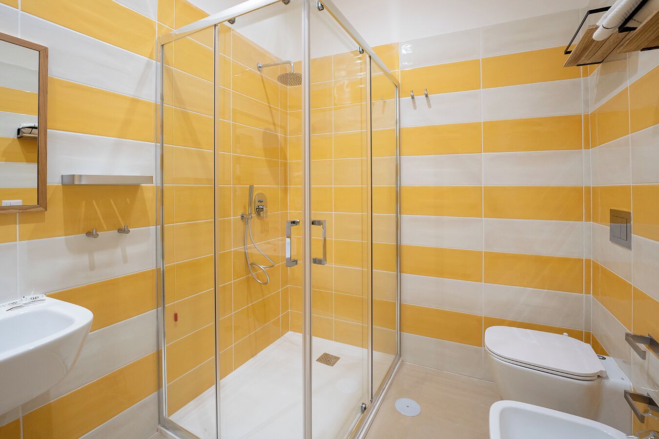 Un bagno con piastrelle giallo e bianco a strisce, una cabina doccia in vetro, un lavandino bianco e un water.