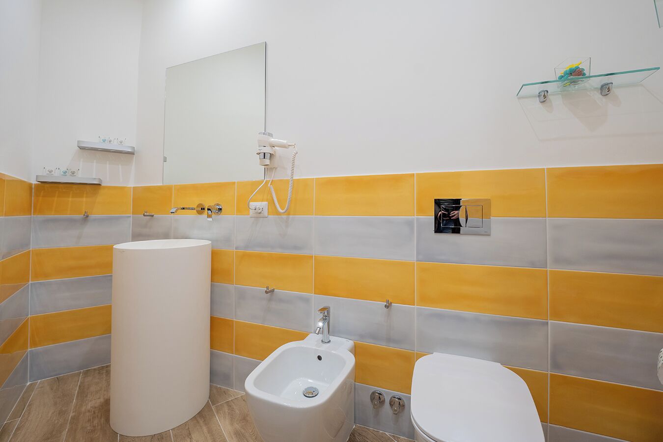 A bathroom with a large white sink, a bidet, and a toilet. There is a mirror above the sink and a shelf with toiletries on the wall.
