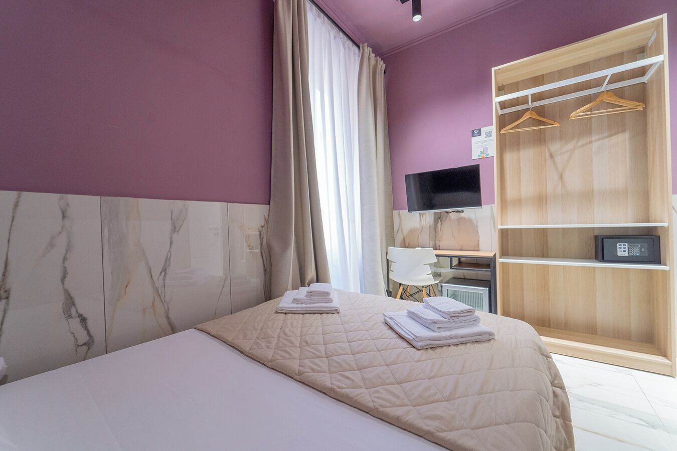 A hotel room with a bed, a desk with a chair, a TV, and a wardrobe with hangers.