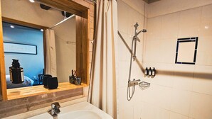 Une salle de bain avec un meuble-lavabo en bois, un grand miroir, une douche équipée d’un pommeau de douche à main et un sol carrelé.