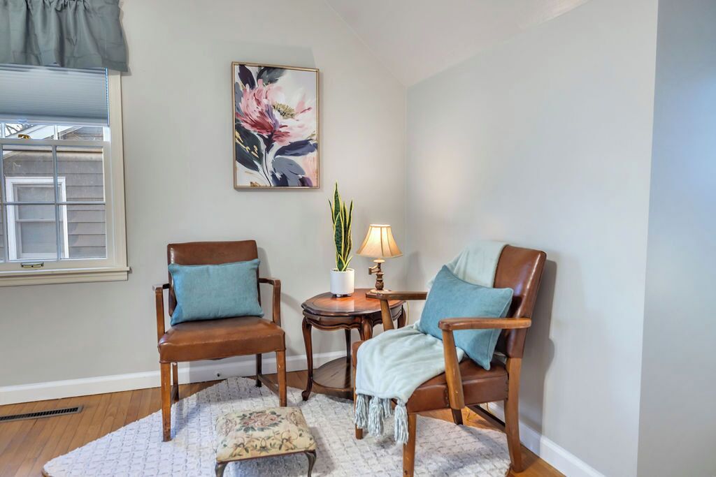 A room with two wooden chairs, a small table with a lamp, a potted plant, and a framed floral artwork on the wall.
