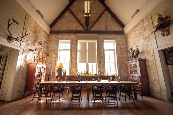 A spacious dining area with a long wooden table and chairs, large windows, stone walls, and various hunting trophies.