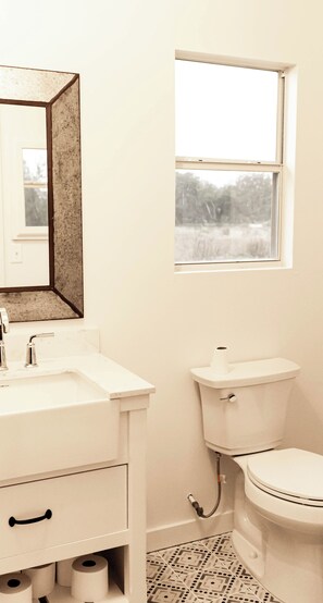 A bathroom with a white sink, mirror, toilet, and a window with a view of trees. - Morris Ranch (Fredericksburg)