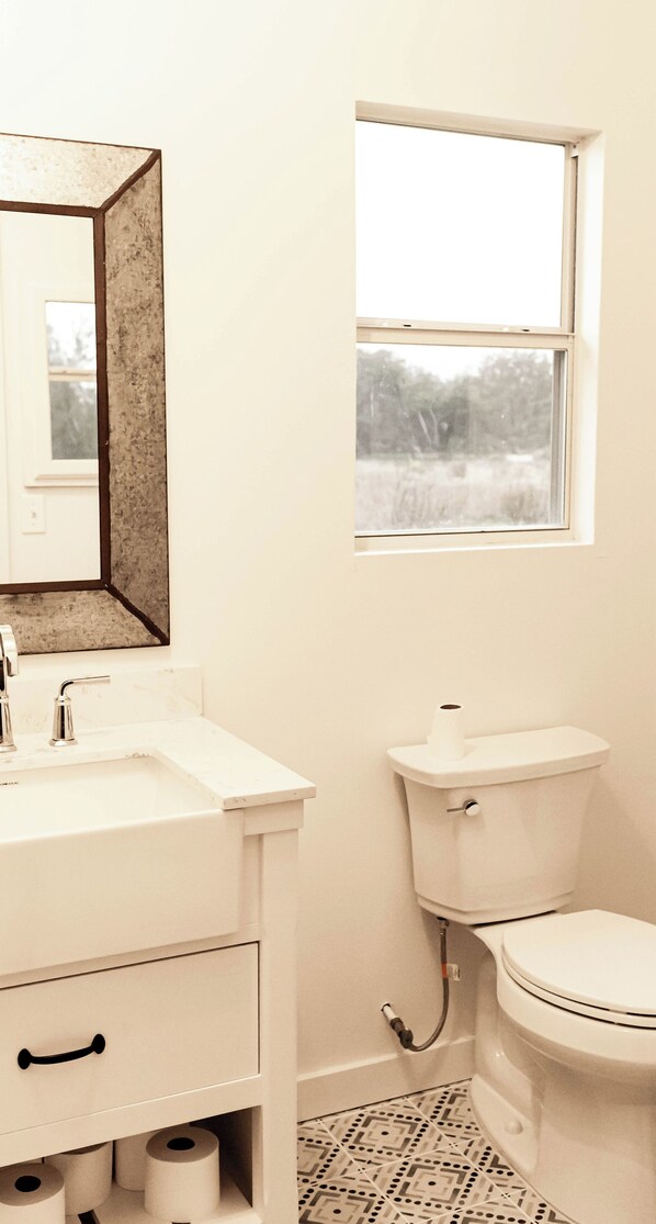 A bathroom with a white sink, mirror, toilet, and a window with a view of trees. - Morris Ranch (Fredericksburg)