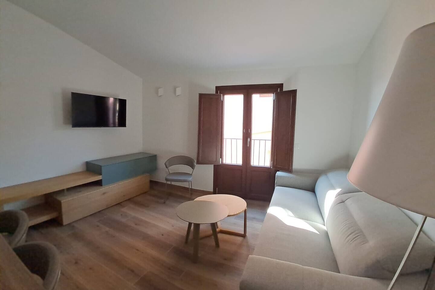 Ein Wohnzimmer mit einer Couch, einem kleinen runden Tisch, einem an der Wand befestigten Fernseher und einem Balkon mit Holztüren.