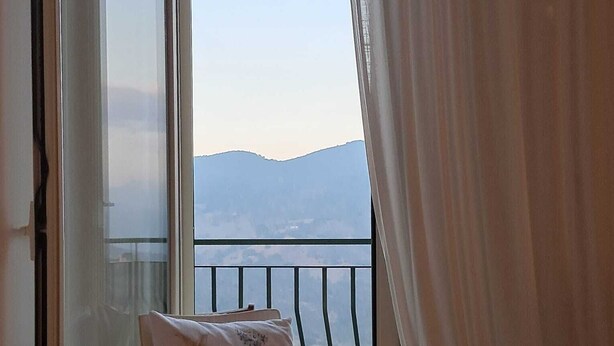Een balkon met uitzicht op verre bergen, een stoel met kussen en lichte gordijnen.