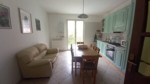 Een woonkamer met een beige bank, een eettafel met stoelen en een keuken met blauwgroene kastjes en een fornuis.