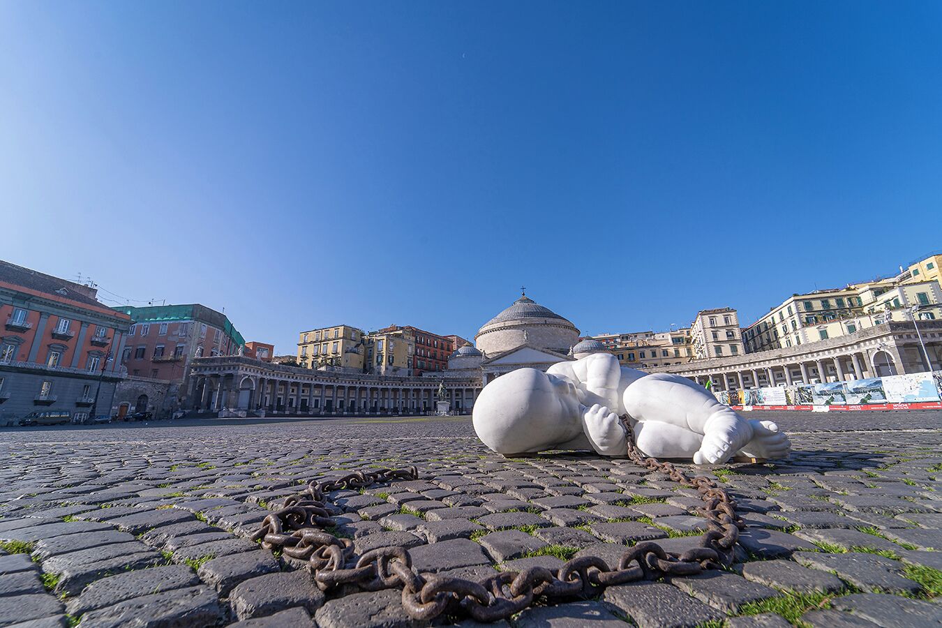 A cobblestone plaza with a large white sculpture of a reclining figure, surrounded by historic buildings and a domed structure in the background.