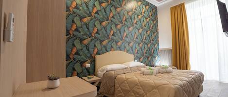 A hotel room with a bed, a desk, a small table with a plant, and a wall with a leaf pattern.