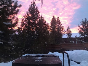 A snowy outdoor area with a wooden bench and a metal table, surrounded by tall evergreen trees and a wooden fence. - Fun-sized 3-bedroom cottage on the river, plus hot tub. Pet-Friendly! (Grand Lake)