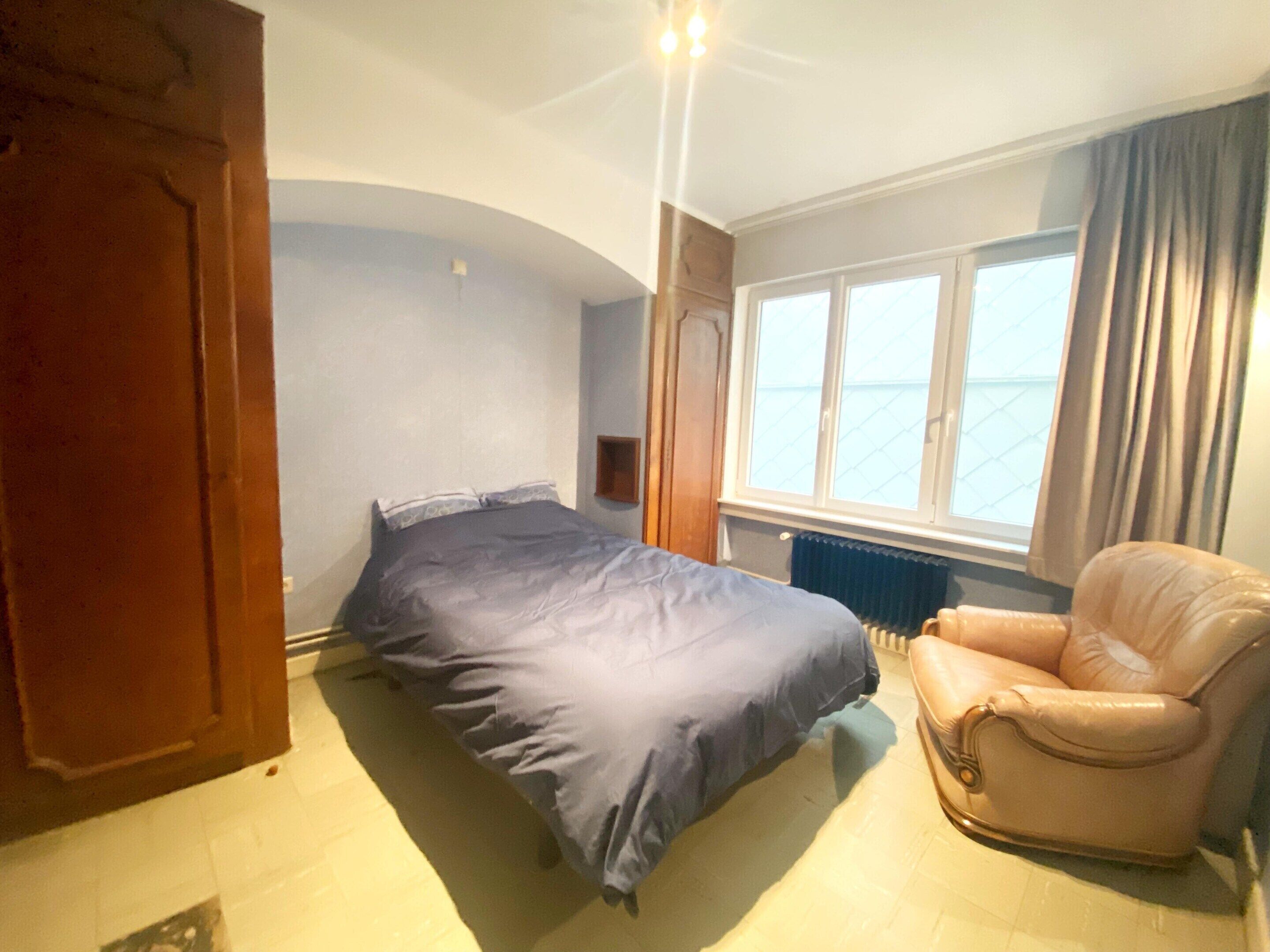 A bedroom with a bed, a chair, a window with curtains, and a radiator.